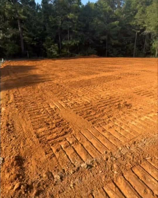Freshly graded red dirt building pad with dozer tracks and pine trees behind it