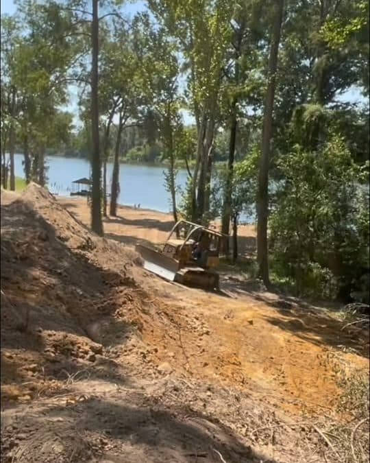 Excavator working red dirt along a wooded lake bank